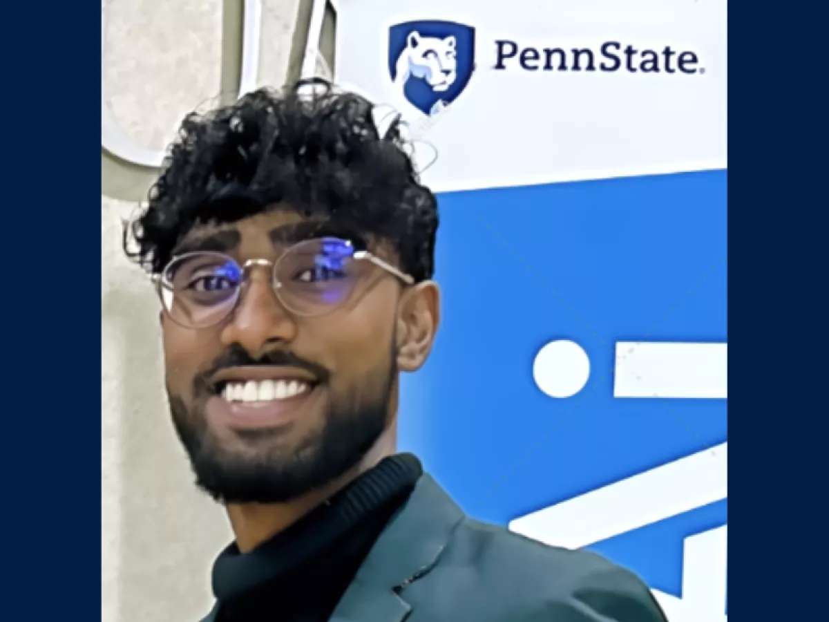 Smiling man with dark hair and glasses standing in front of banner with Penn State logo