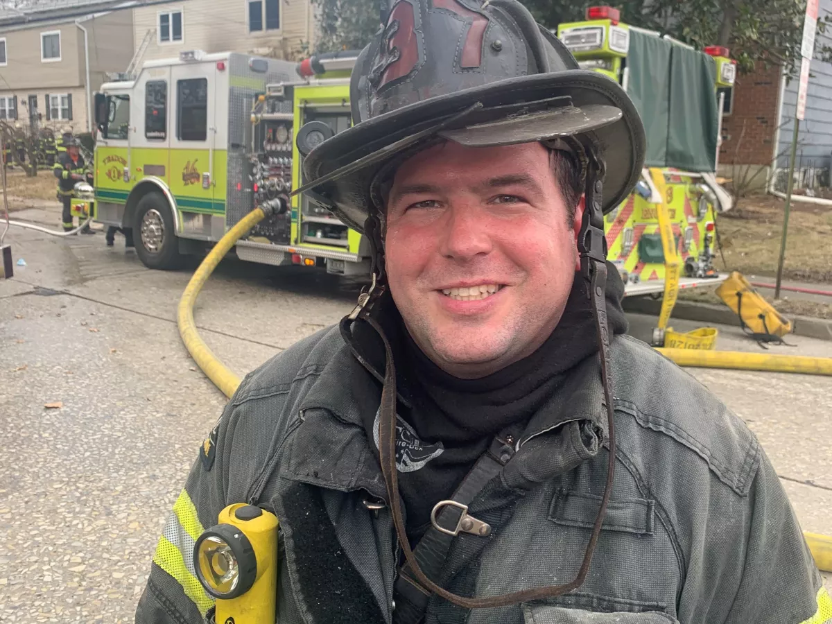 Smiling man wearing firefighter gear with fire truck in background