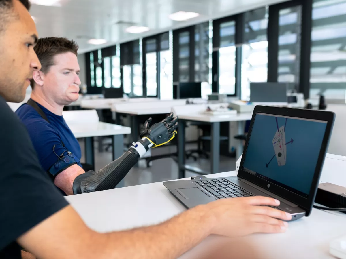 Man sitting in front of laptop, next to man with an attached metal arm.
