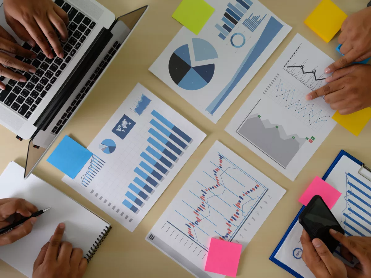 Papers with charts and graphs spread out on table, surrounded by hands using laptop keyboards and phones