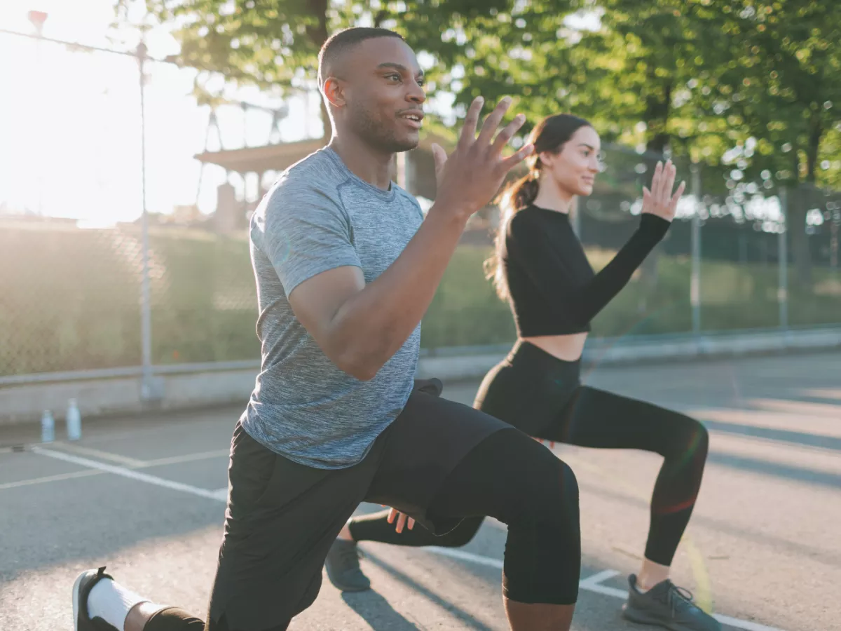 Man and woman in workout attire stretching in a park with trees and sunshine in the background