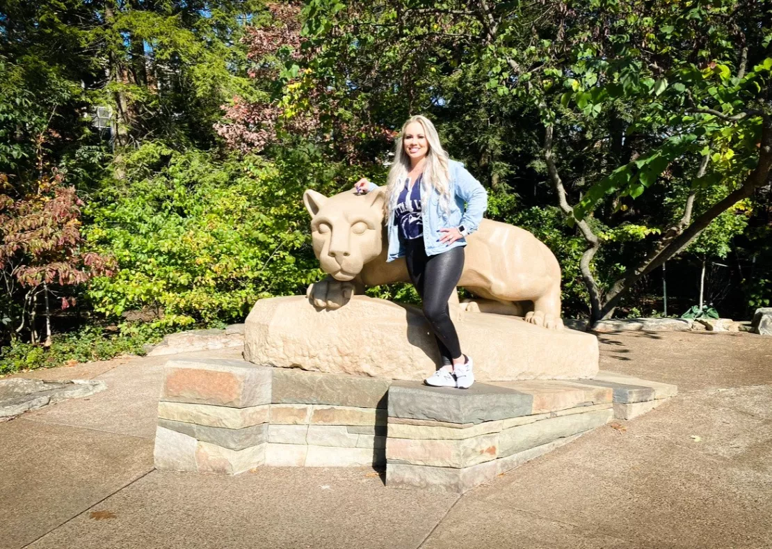 Smiling woman with long blonde hair standing next to the Nittany Lion shrine