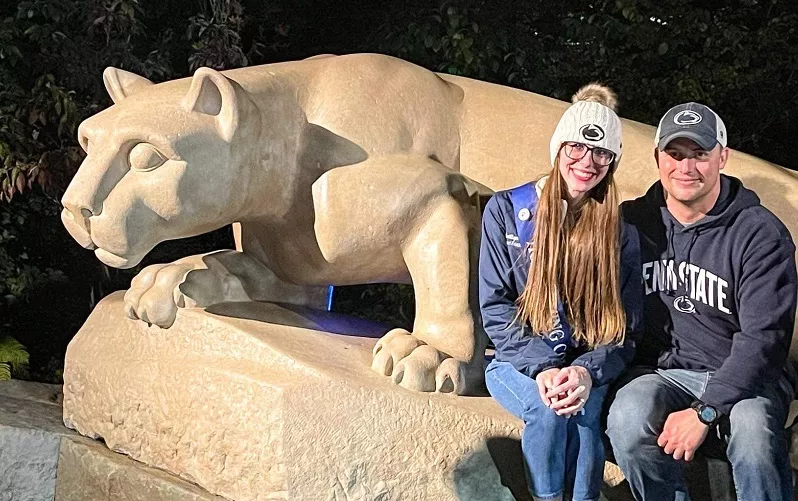 Smiling man and woman in Penn State gear sitting in front of the Nittany Lion shrine